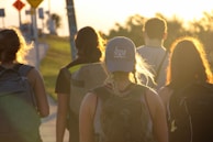 A group of five individuals walking along a path under the warm glow of the setting sun. They carry backpacks, and one wears a cap with text on it. The scenic background includes grassy areas and some roadside signs, evoking a sense of adventure or travel.