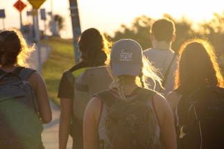 A group of five individuals walking along a path under the warm glow of the setting sun. They carry backpacks, and one wears a cap with text on it. The scenic background includes grassy areas and some roadside signs, evoking a sense of adventure or travel.