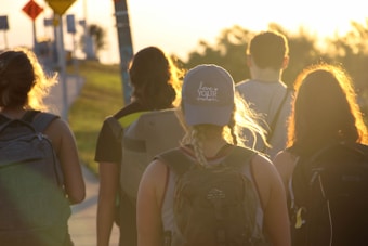 A group of five individuals walking along a path under the warm glow of the setting sun. They carry backpacks, and one wears a cap with text on it. The scenic background includes grassy areas and some roadside signs, evoking a sense of adventure or travel.