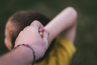A person's hand is holding the hand of a young child. The adult's hand appears to be guiding or leading the child, whose head is visible with short hair and arms positioned behind their head. The focus is primarily on the hands, creating a close-up view with a blurred background.