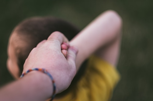 A person's hand is holding the hand of a young child. The adult's hand appears to be guiding or leading the child, whose head is visible with short hair and arms positioned behind their head. The focus is primarily on the hands, creating a close-up view with a blurred background.