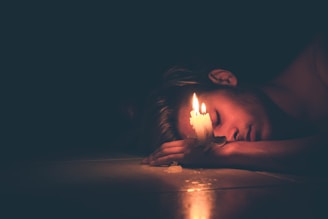A person lies on their side with a lit candle resting on their hand. The warm candlelight softly illuminates the person's face against a dark background, creating a serene and introspective atmosphere.