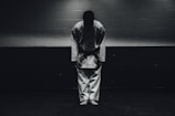 A person dressed in a traditional martial arts uniform stands in a dimly lit room bowing forward, with their feet together and hands by their sides.