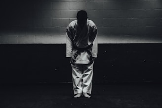 A person dressed in a traditional martial arts uniform stands in a dimly lit room bowing forward, with their feet together and hands by their sides.