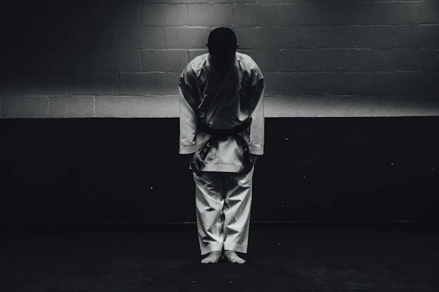 A focused martial artist tying a black sash around their waist in a dimly lit dojo.