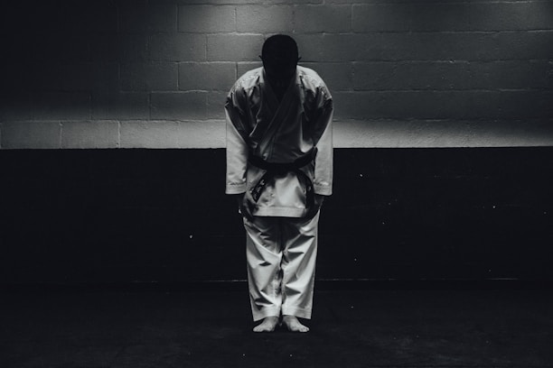 A person dressed in a traditional martial arts uniform stands in a dimly lit room bowing forward, with their feet together and hands by their sides.