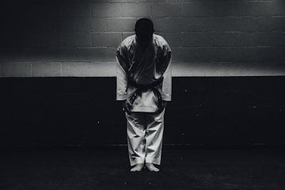 A person dressed in a traditional martial arts uniform stands in a dimly lit room bowing forward, with their feet together and hands by their sides.