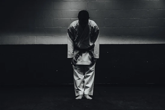 A person dressed in a traditional martial arts uniform stands in a dimly lit room bowing forward, with their feet together and hands by their sides.