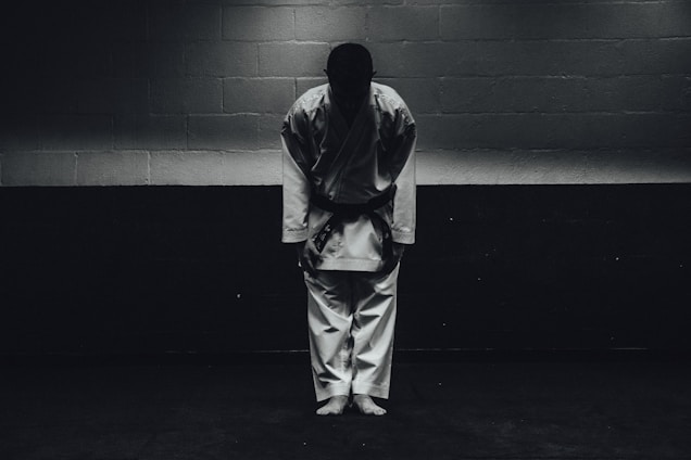 A person dressed in a traditional martial arts uniform stands in a dimly lit room bowing forward, with their feet together and hands by their sides.