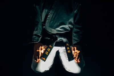 A martial artist is kneeling with their hands placed on their thighs. The black belt around their waist features gold embroidered characters. The lighting is dramatic, highlighting parts of the uniform and creating a sense of focus and intensity.