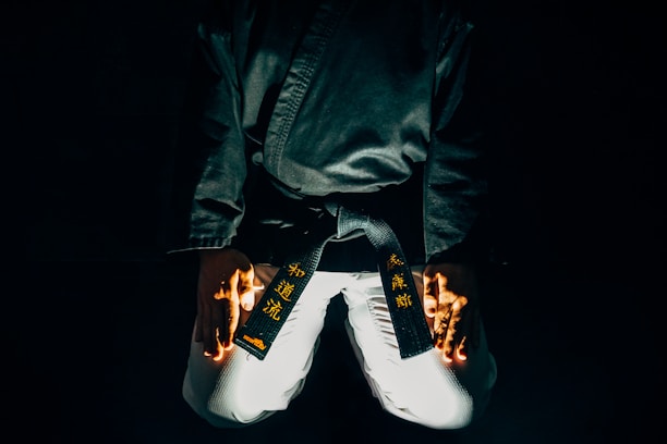 A martial artist is kneeling with their hands placed on their thighs. The black belt around their waist features gold embroidered characters. The lighting is dramatic, highlighting parts of the uniform and creating a sense of focus and intensity.