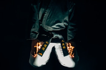 A martial artist is kneeling with their hands placed on their thighs. The black belt around their waist features gold embroidered characters. The lighting is dramatic, highlighting parts of the uniform and creating a sense of focus and intensity.