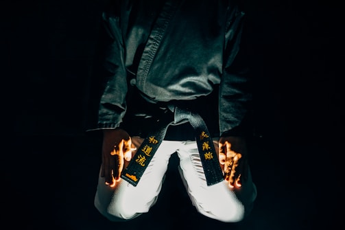 A martial artist is kneeling with their hands placed on their thighs. The black belt around their waist features gold embroidered characters. The lighting is dramatic, highlighting parts of the uniform and creating a sense of focus and intensity.