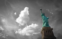 A striking image of the Statue of Liberty standing tall against a dramatic sky filled with fluffy clouds. The statue's vivid green color contrasts with the grayscale background, creating a powerful visual effect.