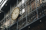 A large, ornate clock is mounted on a metallic, industrial-looking wall structure with a grid of windows. The clock has Roman numerals and detailed design elements, and the setting appears to be inside a historic train station or similar public building.