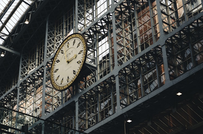 A large, ornate clock is mounted on a metallic, industrial-looking wall structure with a grid of windows. The clock has Roman numerals and detailed design elements, and the setting appears to be inside a historic train station or similar public building.