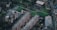An aerial view of an urban neighborhood with rows of brick residential buildings surrounded by roads and green spaces. The scene includes a small sports court, a playground, and a mix of other buildings. Trees and patches of grass add a hint of nature to the cityscape.