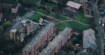 An aerial view of an urban neighborhood with rows of brick residential buildings surrounded by roads and green spaces. The scene includes a small sports court, a playground, and a mix of other buildings. Trees and patches of grass add a hint of nature to the cityscape.