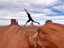 A person is leaping across a gap between two large red rock formations in a desert landscape. The background includes expansive views of similar mesas and buttes under a cloudy blue sky.