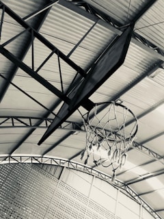 A basketball hoop with a frayed net is attached to a metal backboard, hanging from an indoor ceiling with a corrugated metal roof structure. The perspective is looking upwards, capturing the roof's beams and metal grating on the walls.