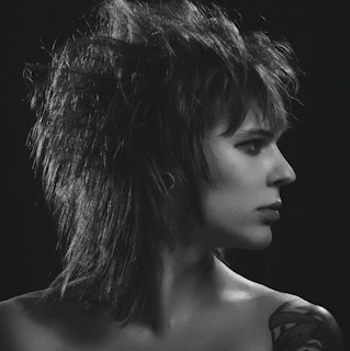 A black and white portrait of a person with striking, spiky hair styled in a punk fashion, facing to the right. The individual has a neutral expression and a visible tattoo on the shoulder. The lighting highlights the intricate details of the hair and facial features against a dark background.