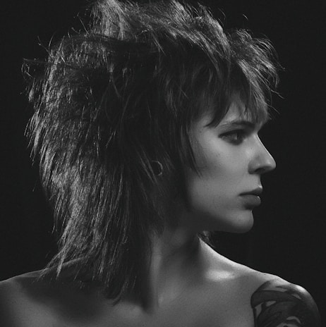 A black and white portrait of a person with striking, spiky hair styled in a punk fashion, facing to the right. The individual has a neutral expression and a visible tattoo on the shoulder. The lighting highlights the intricate details of the hair and facial features against a dark background.