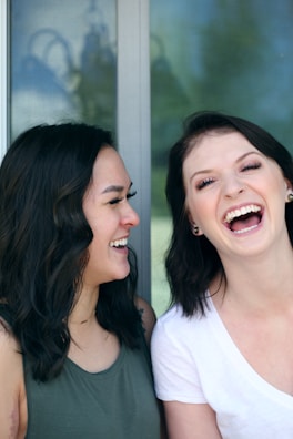 Two women with dark hair are laughing and smiling warmly at each other. One is wearing a dark green sleeveless top while the other is in a white t-shirt. The background is a window with a faint reflection visible.