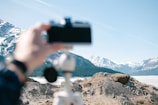 A hand is holding a camera in the foreground, capturing a scenic view of a mountainous landscape with snow-covered peaks in the distance. The terrain is rocky and rugged, and a tripod is visible in the foreground. The sky above is clear and blue.