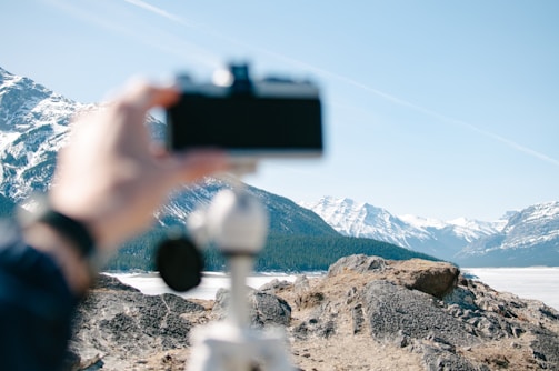 A hand is holding a camera in the foreground, capturing a scenic view of a mountainous landscape with snow-covered peaks in the distance. The terrain is rocky and rugged, and a tripod is visible in the foreground. The sky above is clear and blue.