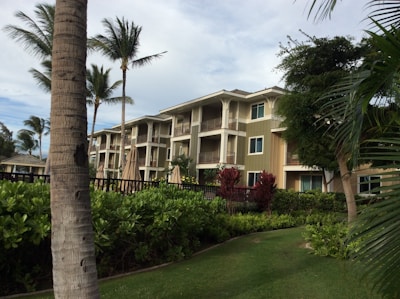 A three-story residential building is surrounded by lush tropical foliage and palm trees. The architecture features a mix of wood and neutral colors with balconies on each floor. The landscape is well-maintained with green grass and various types of plants. A clear sky with some clouds is visible above.