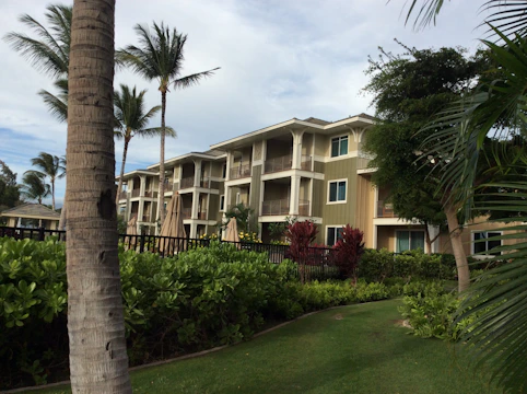 A three-story residential building is surrounded by lush tropical foliage and palm trees. The architecture features a mix of wood and neutral colors with balconies on each floor. The landscape is well-maintained with green grass and various types of plants. A clear sky with some clouds is visible above.