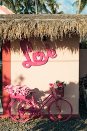 A pink bicycle is adorned with vibrant flowers in its front and rear baskets, standing against a beige wall with a large pink 'Love' sign. The building has a thatched roof, and palm trees can be seen in the background, suggesting a tropical setting.