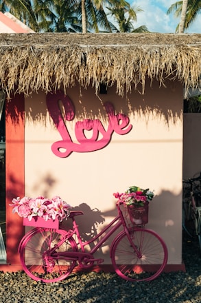 A pink bicycle is adorned with vibrant flowers in its front and rear baskets, standing against a beige wall with a large pink 'Love' sign. The building has a thatched roof, and palm trees can be seen in the background, suggesting a tropical setting.