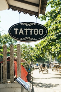 A vintage-style sign with the word 'Tattoo' hangs prominently over a wooden fence. The background features a sunny street scene with trees providing shade, and a busy sidewalk where people are moving about. Some structures with red awnings and carts are visible in the distance, giving a quaint and rustic feel.