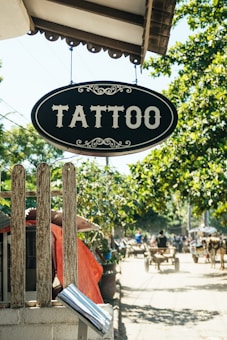 A vintage-style sign with the word 'Tattoo' hangs prominently over a wooden fence. The background features a sunny street scene with trees providing shade, and a busy sidewalk where people are moving about. Some structures with red awnings and carts are visible in the distance, giving a quaint and rustic feel.
