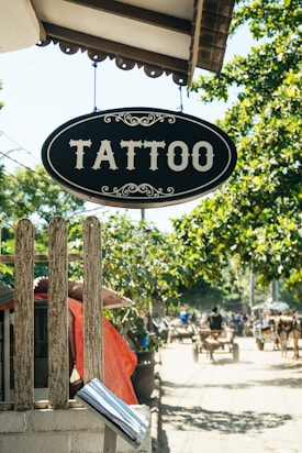 A vintage-style sign with the word 'Tattoo' hangs prominently over a wooden fence. The background features a sunny street scene with trees providing shade, and a busy sidewalk where people are moving about. Some structures with red awnings and carts are visible in the distance, giving a quaint and rustic feel.