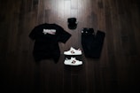 A neatly arranged flat lay featuring a black Balenciaga t-shirt, black jeans, a black baseball cap, and a pair of white sneakers with red and green accents. The items are placed on a dark wooden floor, creating a stylish and modern aesthetic.