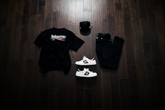 A neatly arranged flat lay featuring a black Balenciaga t-shirt, black jeans, a black baseball cap, and a pair of white sneakers with red and green accents. The items are placed on a dark wooden floor, creating a stylish and modern aesthetic.