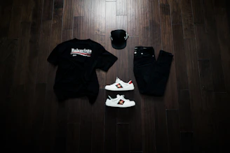Flat lay of solid black and off-white t-shirts with red tags, styled with streetwear accessories on a concrete floor.