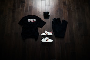A neatly arranged flat lay featuring a black Balenciaga t-shirt, black jeans, a black baseball cap, and a pair of white sneakers with red and green accents. The items are placed on a dark wooden floor, creating a stylish and modern aesthetic.
