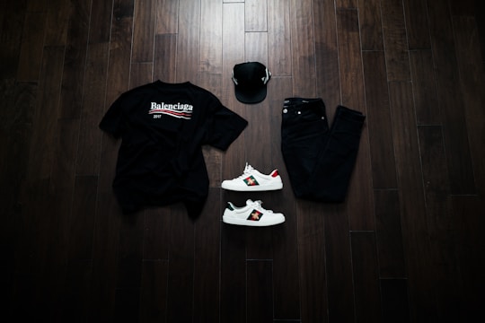 A neatly arranged flat lay featuring a black Balenciaga t-shirt, black jeans, a black baseball cap, and a pair of white sneakers with red and green accents. The items are placed on a dark wooden floor, creating a stylish and modern aesthetic.