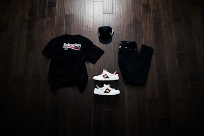 A neatly arranged flat lay featuring a black Balenciaga t-shirt, black jeans, a black baseball cap, and a pair of white sneakers with red and green accents. The items are placed on a dark wooden floor, creating a stylish and modern aesthetic.