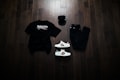 A neatly arranged flat lay featuring a black Balenciaga t-shirt, black jeans, a black baseball cap, and a pair of white sneakers with red and green accents. The items are placed on a dark wooden floor, creating a stylish and modern aesthetic.