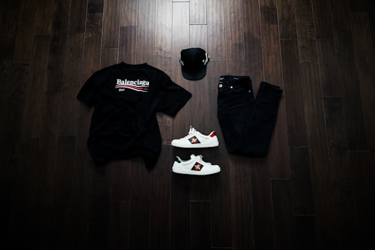A neatly arranged flat lay featuring a black Balenciaga t-shirt, black jeans, a black baseball cap, and a pair of white sneakers with red and green accents. The items are placed on a dark wooden floor, creating a stylish and modern aesthetic.