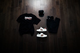 A neatly arranged flat lay featuring a black Balenciaga t-shirt, black jeans, a black baseball cap, and a pair of white sneakers with red and green accents. The items are placed on a dark wooden floor, creating a stylish and modern aesthetic.