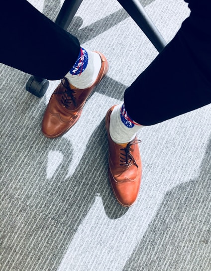 A pair of brown leather dress shoes is worn with white socks that have a blue and red pattern. The person is seated, and the floor is covered with a textured gray carpet, partly obscured by the shadows of a chair’s legs.