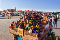 A vibrant market scene features a stall filled with colorful ceramic tagines and traditional pottery. Various stalls and people are visible in the bustling background, with a historic tower and buildings providing a scenic backdrop. Red tents and awnings add to the lively atmosphere.