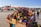 A vibrant market scene features a stall filled with colorful ceramic tagines and traditional pottery. Various stalls and people are visible in the bustling background, with a historic tower and buildings providing a scenic backdrop. Red tents and awnings add to the lively atmosphere.