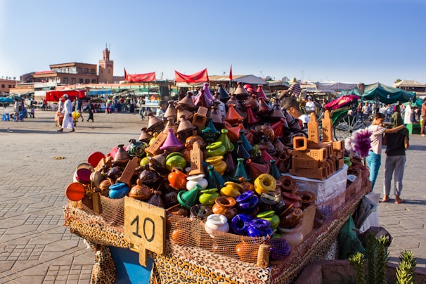 A vibrant market scene features a stall filled with colorful ceramic tagines and traditional pottery. Various stalls and people are visible in the bustling background, with a historic tower and buildings providing a scenic backdrop. Red tents and awnings add to the lively atmosphere.