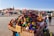 A vibrant market scene features a stall filled with colorful ceramic tagines and traditional pottery. Various stalls and people are visible in the bustling background, with a historic tower and buildings providing a scenic backdrop. Red tents and awnings add to the lively atmosphere.